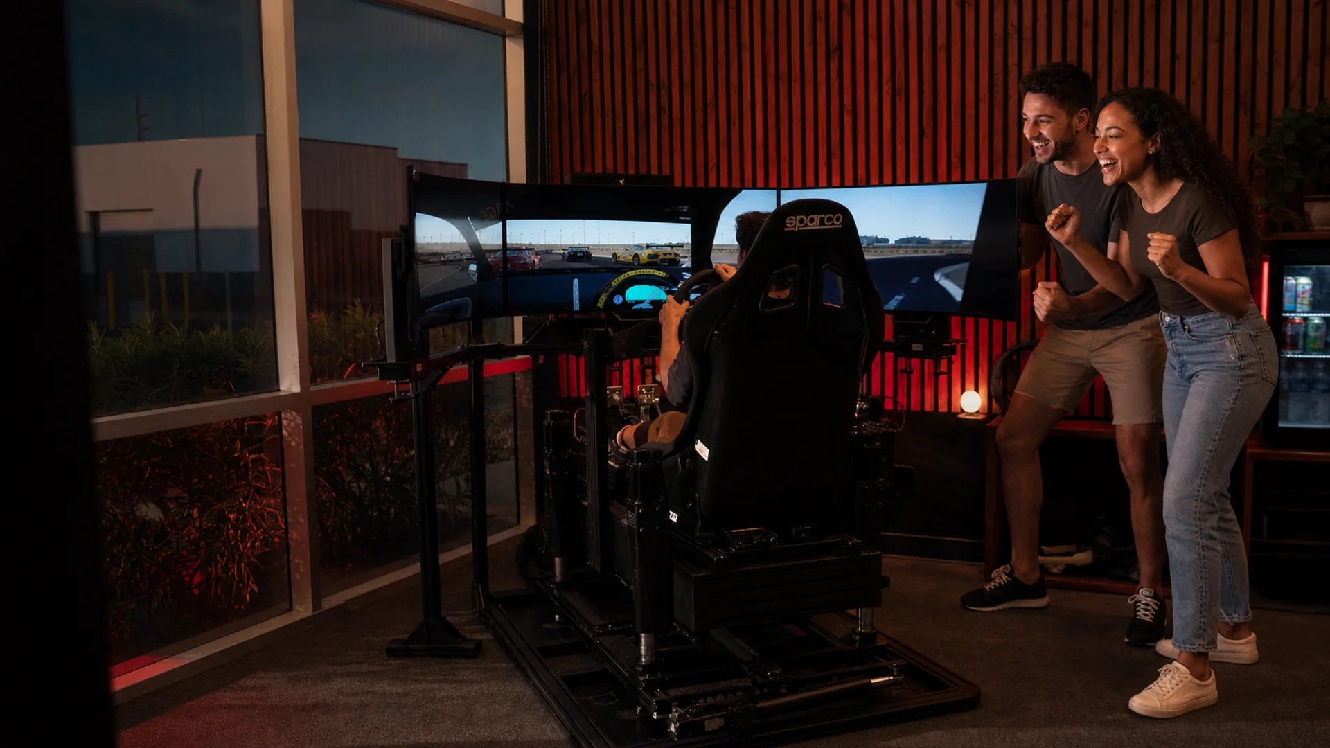 Adults cheer and smile around a professional racing simulator at a birthday event